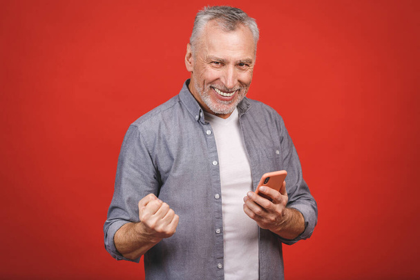 Buon uomo anziano maturo in casual utilizzando smartphone e rendendo il gesto vincitore isolato su sfondo rosso. Uso del telefono
. - Foto, immagini