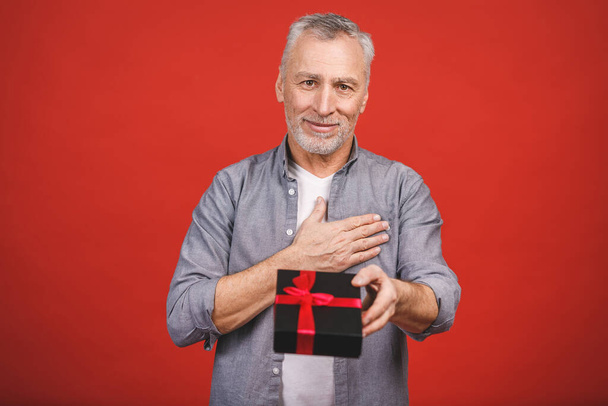 Mature senior man showing a gift with red ribbon as a present isolated against red background. - 写真・画像