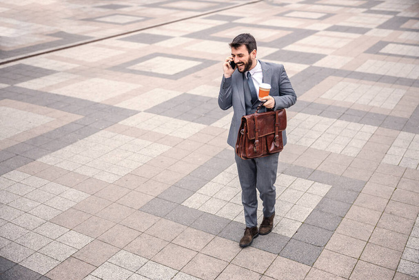 Jeune homme d'affaires barbu souriant tenant son sac et son café du matin tout en allant au travail et avoir une conversation téléphonique
. - Photo, image