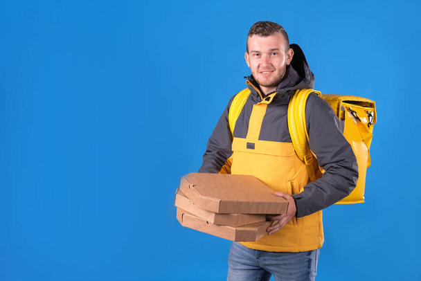 Happy food delivery man holds pizzas in cardboard boxes and holds them out for customer, dressed in yellow uniform and refrigerator bag on his back on blue background. Courier food delivery to home. - Photo, image