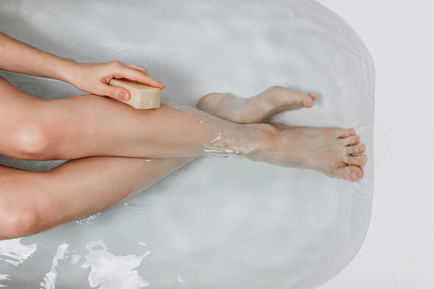 Flatlay of woman 's legs in a bath holding natural homemade mýdlo in one hand - Fotografie, Obrázek