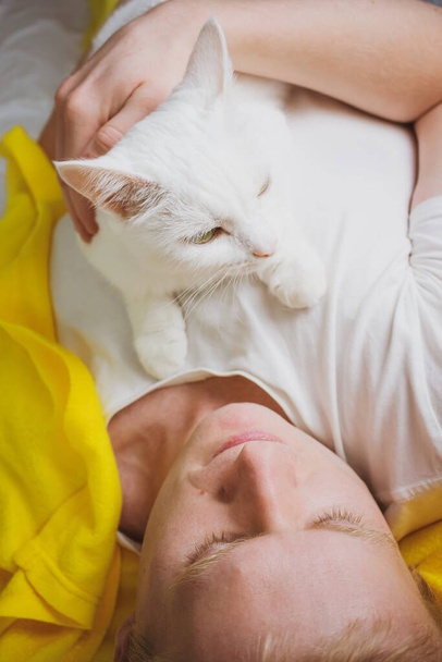 A young blond man is lying on a bed with a white shorthair cat. The concept of pets, weekends, laziness, morning. Photo in bright white and yellow tones. - Photo, Image