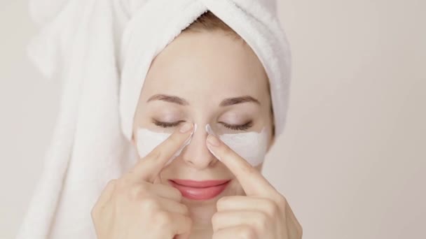 Portrait of beautiful Caucasian young woman with hair wrapped in white towel, applying facial clay mask touching her face. Beauty Treatments. Dayspa. - Filmagem, Vídeo