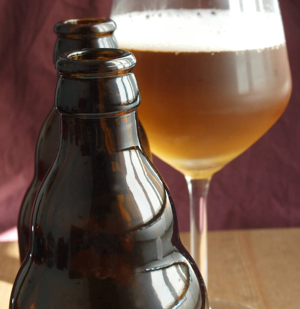 duas garrafas de cerveja de vidro marrom e uma xícara de cerveja sobre fundo de tecido escuro
    - Foto, Imagem