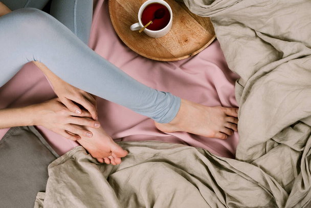 Cozy flatlay of woman's legs in blue leggins in bed with cup of tea, selective focus - Zdjęcie, obraz