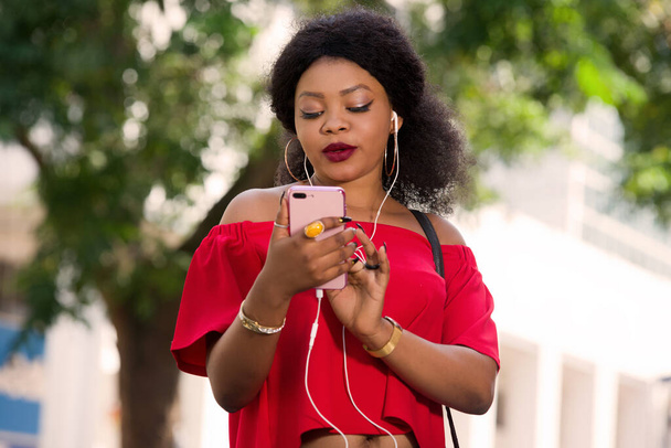Glückliche junge Frau in rotem T-Shirt mit Kopfhörer, die in der Stadt mit dem Handy telefoniert - Foto, Bild