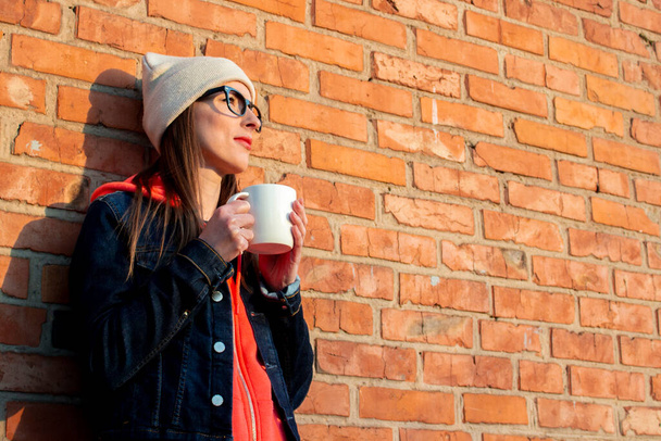 Jonge glimlachende vrouw die wegkijkt in een bril met een witte beker op de achtergrond van een bakstenen muur met een grijze pet, hoodie en spijkerjasje. - Foto, afbeelding