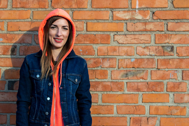 フードを着た若い女性が赤いレンガの壁を背景にデニムジャケットを着ている. - 写真・画像