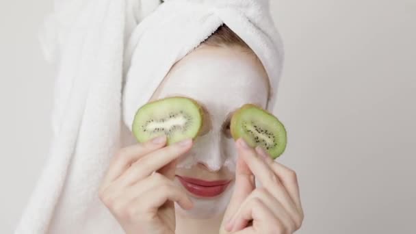 Close up of a girl with white mask on her face and a towel on her head closing her eyes with kiwi fruit slices and smiling. Face Skin Care Concept - Video