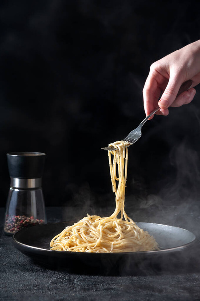 Cacio e Pepe - Pasta italiana picante con queso y pimienta en la placa negra, mujer sosteniendo espaguetis tenedor sobre fondo oscuro
 - Foto, imagen