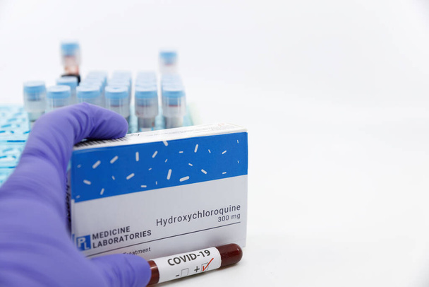 Dubai-UAE-Circa 2020:Doctor showing box of medicine with positive covid-19 test.Concept of Hydroxychloroquine medicine with blood tests tubes on the background.Cure for coronavirus,COVID-19 treatment. - Фото, зображення