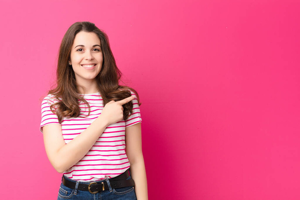 young pretty woman smiling cheerfully, feeling happy and pointing to the side and upwards, showing object in copy space against pink wall - Foto, Imagen