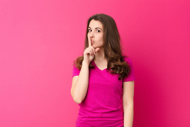 young pretty woman asking for silence and quiet, gesturing with finger in front of mouth, saying shh or keeping a secret against pink wall - Photo, Image