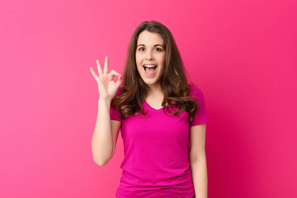junge hübsche Frau, die sich erfolgreich und zufrieden fühlt, mit weit geöffnetem Mund lächelt, mit der Hand Okay-Zeichen gegen rosa Wand macht - Foto, Bild