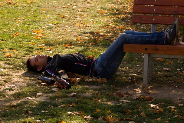 Пьяный молодой человек, спящий днем на траве под скамейкой. Проблема алкоголизма среди молодежи. - Фото, изображение
