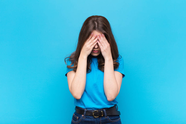 young pretty woman feeling sad, frustrated, nervous and depressed, covering face with both hands, crying against blue wall - Fotoğraf, Görsel