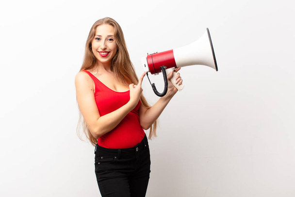 yound blonde woman smiling cheerfully, feeling happy and pointing to the side and upwards, showing object in copy space holding a megaphone - Fotoğraf, Görsel