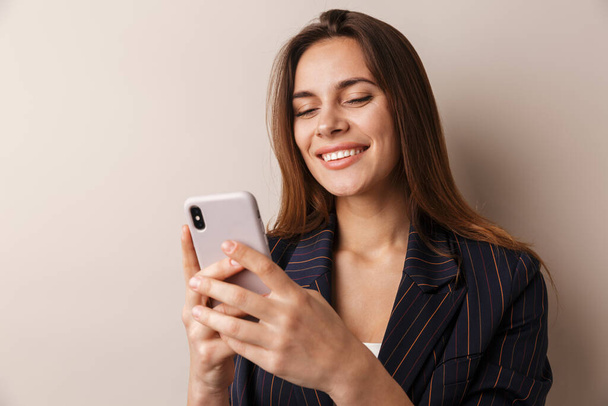 Photo of cheerful businesswoman in formal suit laughing and using cellphone isolated over white background - Valokuva, kuva