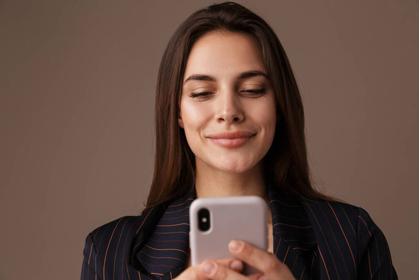 Photo of pleased businesswoman in formal suit smiling and using cellphone isolated over grey background - Valokuva, kuva