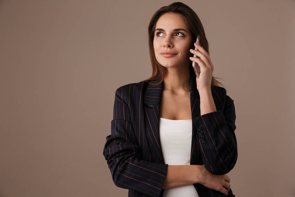 Photo of pleased businesswoman in formal suit smiling and talking on cellphone isolated over grey background - Photo, image