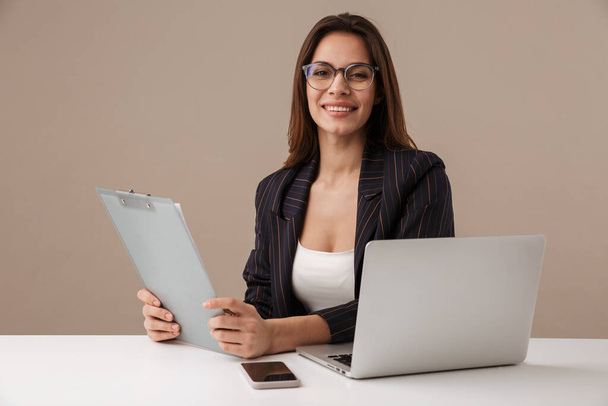 Photo of smiling businesswoman holding clipboard while working with laptop isolated over grey background - Photo, Image
