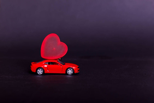 Valentine's day on dark background with miniature red toy car carrying a heart on black surface. Empty place for your text. - Photo, Image