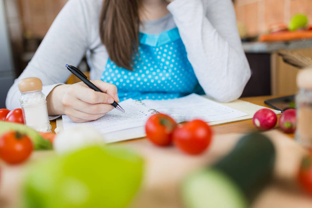 Close up of woman in apron sitting at kitchen table and writing vegetable meal recipe. - Foto, imagen
