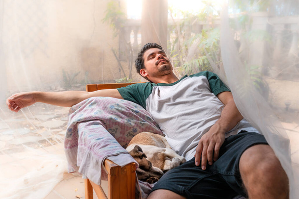 Młody człowiek śpi w fotelu w ogrodzie pod moskitierą. obok niego śpi pies. - Zdjęcie, obraz