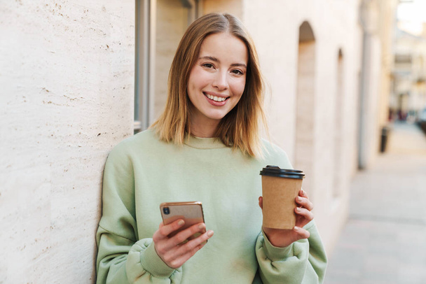 Portrait of smiling young woman drinking coffee takeaway and using mobile phone while walking at city street - Foto, Imagen