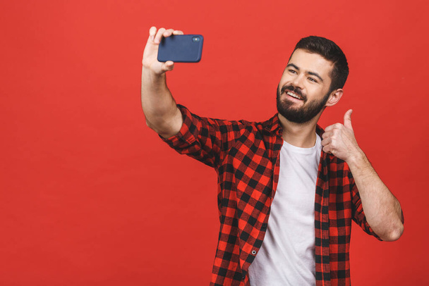 Κοντινό πορτραίτο ενός χαρούμενου γενειοφόρου άντρα που βγάζει selfie πάνω από κόκκινο φόντο. Αντίχειρες επάνω. - Φωτογραφία, εικόνα