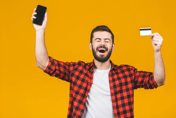 Portrait of a smiling young man holding blank screen mobile phone and showing credit card isolated over yellow background. - Photo, Image