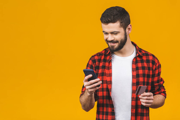 Cheerful young man wearing plaid shirt standing isolated over yellow background, holding mobile phone, showing plastic credit card. - Photo, image