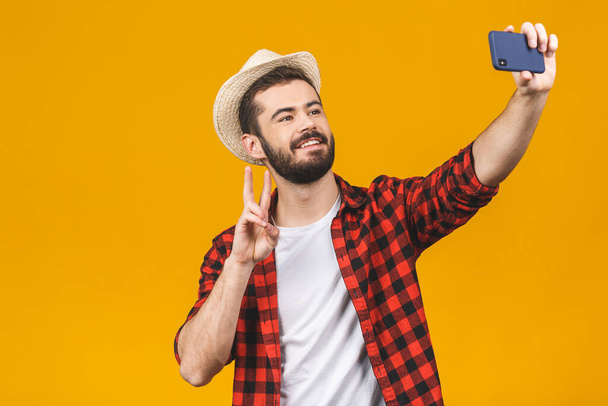 Χαρούμενος νεαρός άνδρας ταξίδια στο καπέλο λήψη selfie με smartphone απομονώνονται σε κίτρινο φόντο. - Φωτογραφία, εικόνα