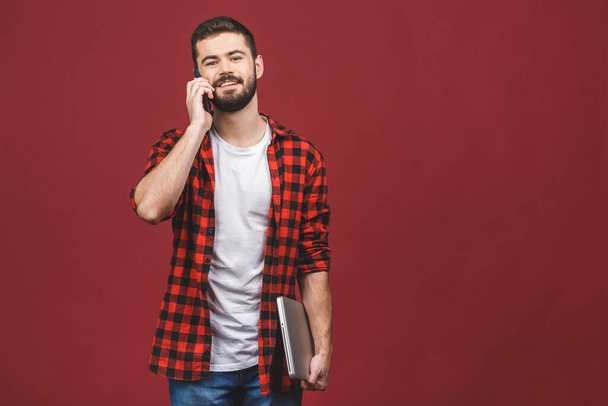 Portrait d'un homme d'affaires souriant heureux dans un ordinateur portable tenue occasionnelle tout en se tenant debout et en parlant sur un téléphone mobile isolé sur fond rouge
. - Photo, image