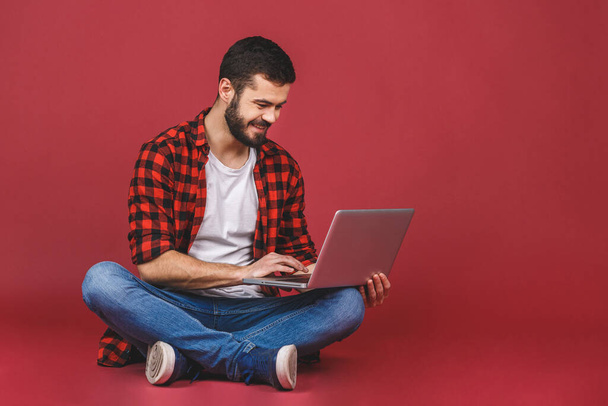 Portret szczęśliwego młodzieńca w niezobowiązującym laptopie, siedzącego na podłodze odizolowanego na czerwonym tle. - Zdjęcie, obraz