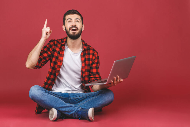 Портрет счастливого молодого человека в белой футболке с ноутбуком, сидящего на полу и указывающего пальцем на красный фон
. - Фото, изображение