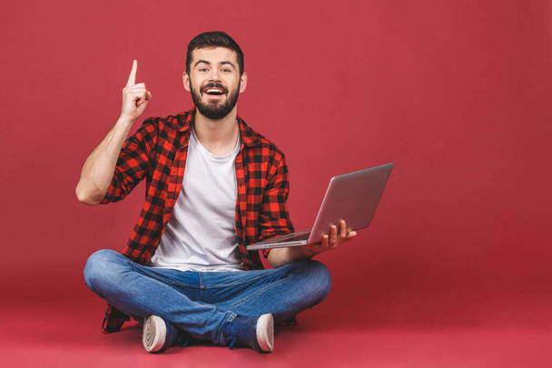 Portrét šťastného mladého muže v bílém tričku držícího notebook, zatímco sedí na podlaze a ukazuje prstem nahoru, izolovaný nad červeným pozadím. - Fotografie, Obrázek