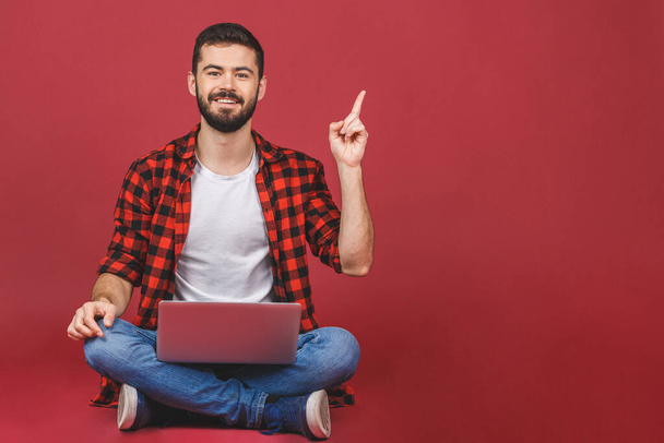 Portrait of a happy young man in casual holding laptop computer while sitting on a floor and pointing finger up isolated over red background. - Photo, Image