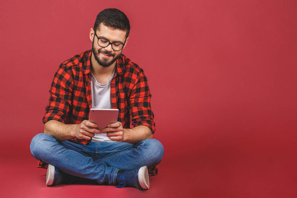 Portret van een jongeman die op de vloer zit met een tablet pc, geïsoleerd tegen een rode achtergrond. Klaar voor uw ontwerp. - Foto, afbeelding