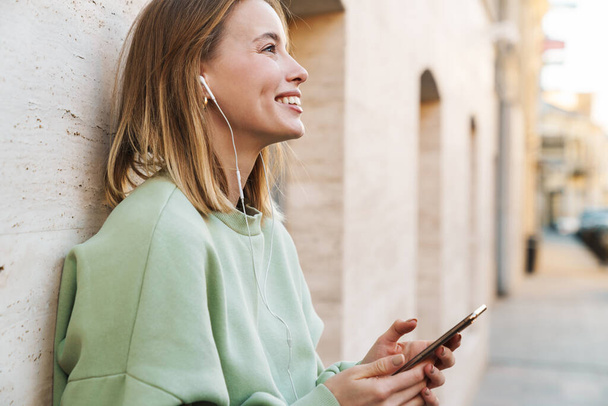Portrait of smiling young woman using earphones and mobile phone while walking at city street - Fotoğraf, Görsel