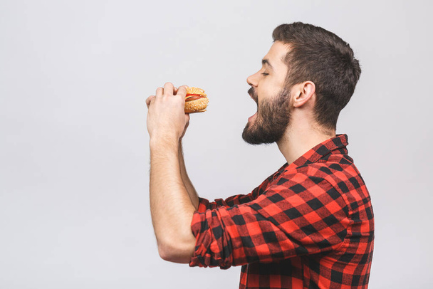 Young man holding a piece of hamburger. Student eats fast food. Burger is not helpful food. Very hungry guy. Diet concept isolated against white background.  - Zdjęcie, obraz