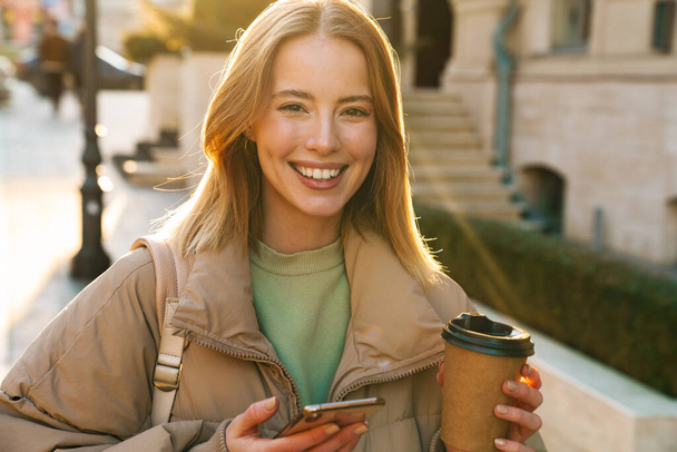 Portrait of joyful caucasian woman using cellphone and drinking coffee takeaway while walking at city street - Photo, Image