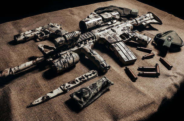 Photo d'un fusil militaire AR-15 camouflé avec lunette de visée posée avec couteau, pistolet et gants tactiques vue de côté
. - Photo, image
