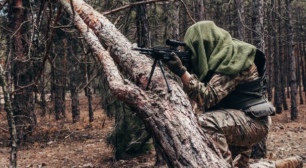 Foto van een volledig uitgeruste gecamoufleerde bossluipschutter met geweer zittend en richtend bij de boom in het bos. - Foto, afbeelding