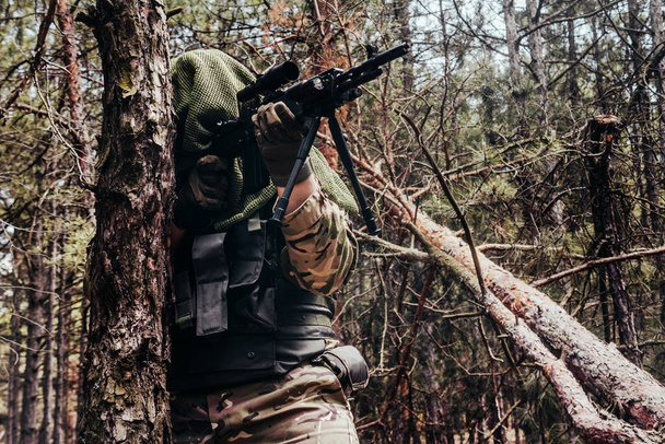 Foto de un francotirador forestal camuflado totalmente equipado con rifle de pie y apuntando en maderas
. - Foto, Imagen