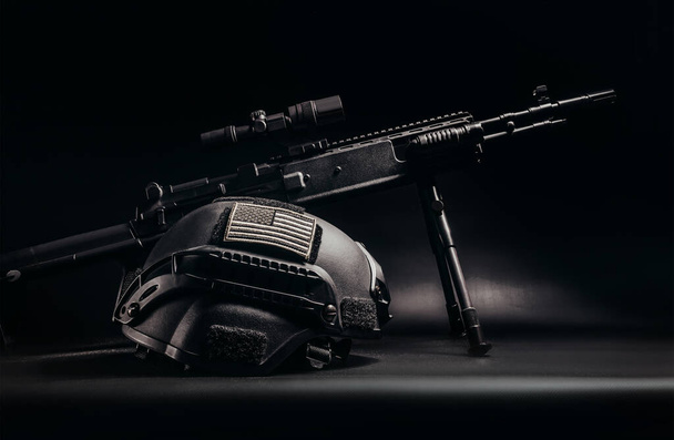 Foto de um rifle sniper preto em pé na mesa com capacete soldado com patch bandeira americana na vista lateral de fundo preto
. - Foto, Imagem