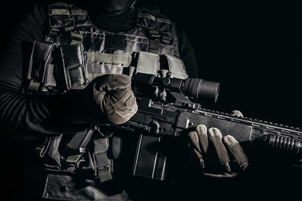 Foto van een soldaat in tactische handschoenen en pantser vest staande en herladen sluipschutter geweer op zwarte achtergrond. - Foto, afbeelding