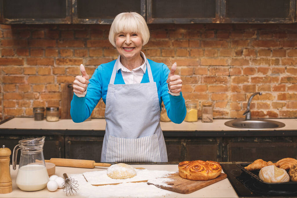 Egy vonzó idős nő portréja főz a konyhában. Nagymama finom süteményt süt. Hüvelykujj fel!. - Fotó, kép