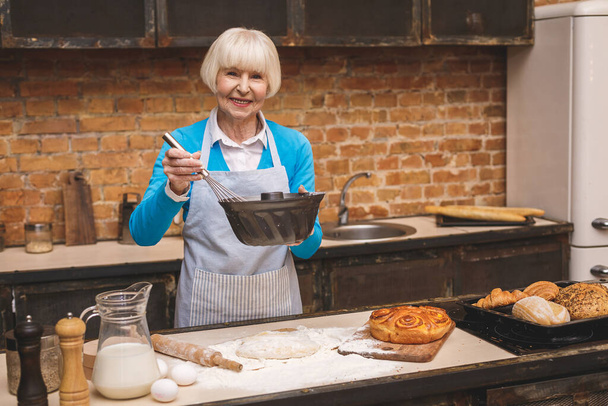 Πορτρέτο της ελκυστική χαμογελαστή ευτυχισμένη ηλικιωμένη γυναίκα μαγειρεύει στην κουζίνα. Η γιαγιά φτιάχνει νόστιμο ψήσιμο..  - Φωτογραφία, εικόνα