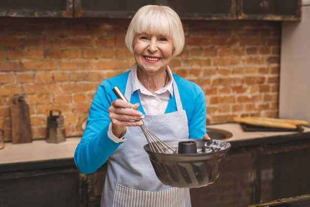Portret van een aantrekkelijke glimlachende oudere vrouw kookt in de keuken. Grootmoeder maakt smakelijk bakken.  - Foto, afbeelding